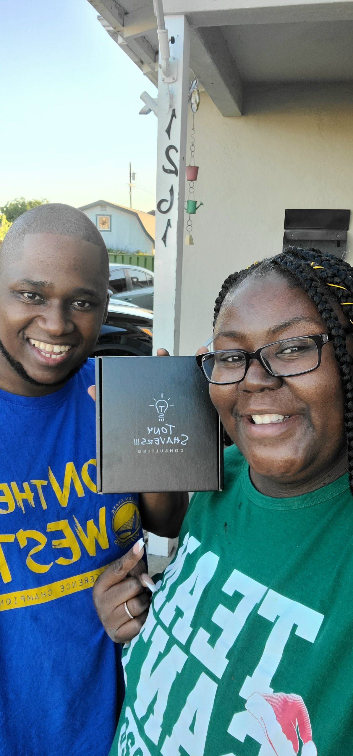 A handsome, young couple, smiling and standing on the side of their home while holding their newly arrived Press Reset affirmation card deck.