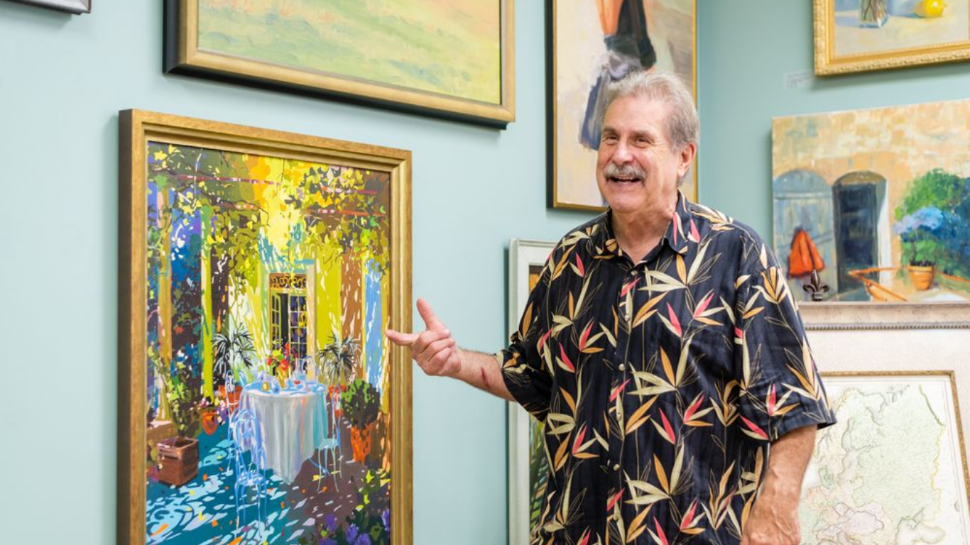 Man standing in an art gallery beside a colorful abstract painting, gesturing toward the artwork.