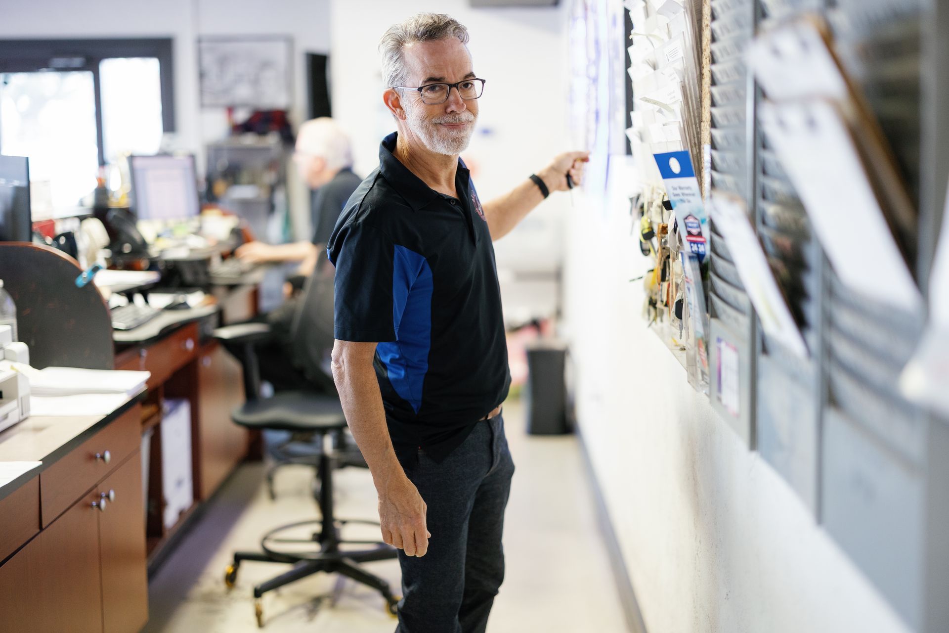 Man pointing at items on a wall, indoors. He wears glasses and a black and blue shirt.
