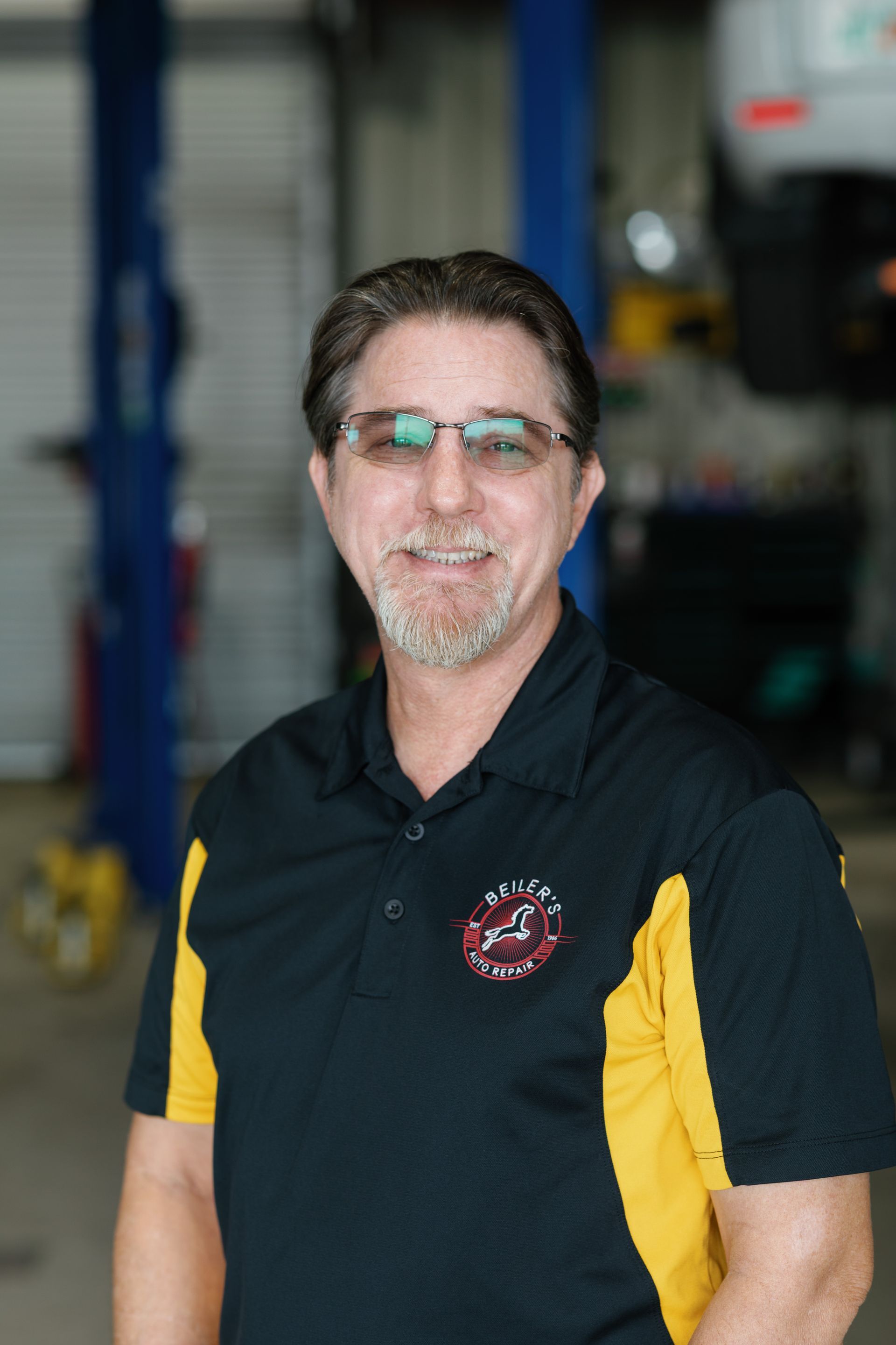 Man wearing glasses, smiling, in a black and yellow polo shirt, standing in a car repair shop.