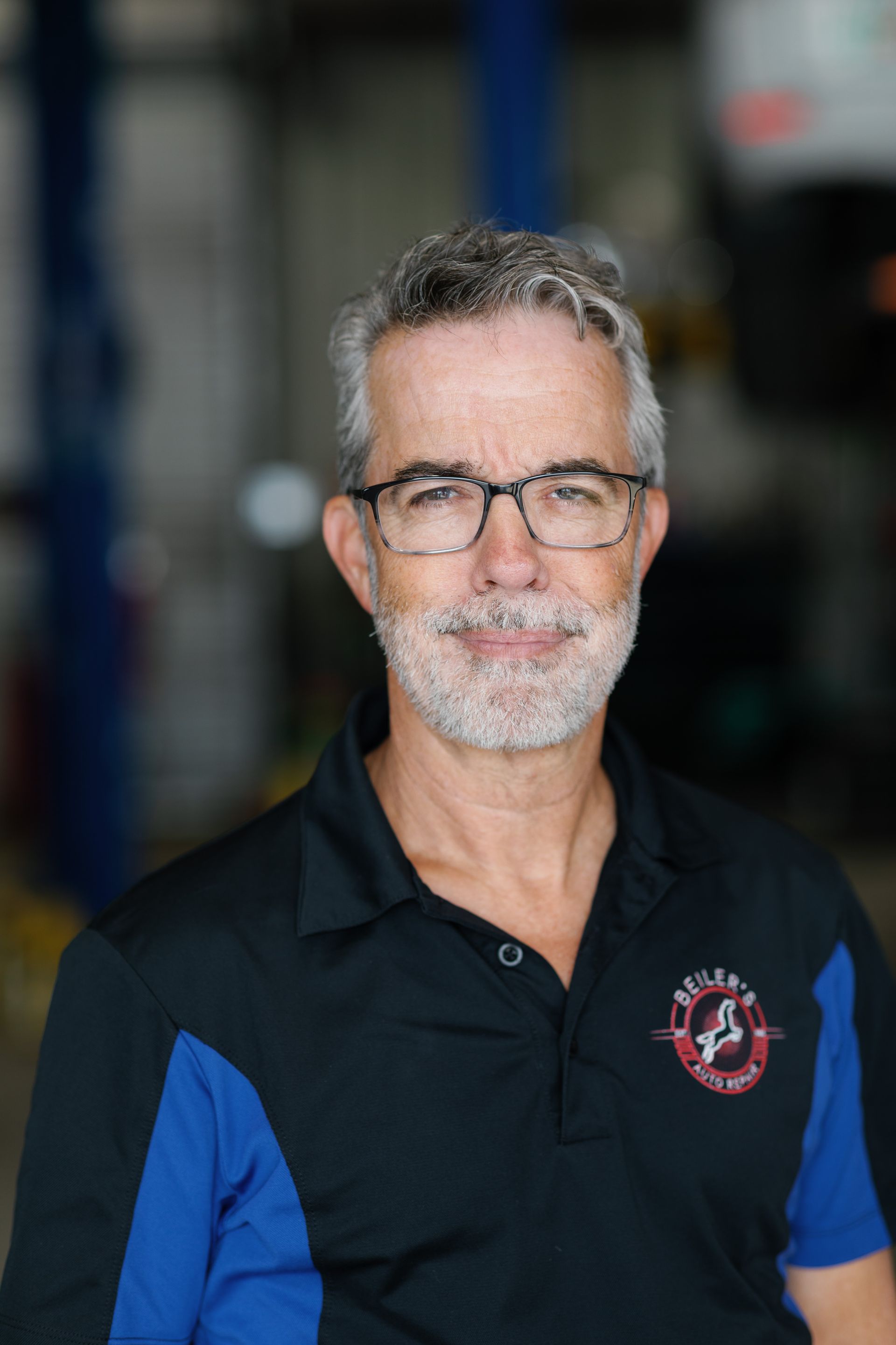 Man wearing glasses and polo shirt, standing in auto shop.