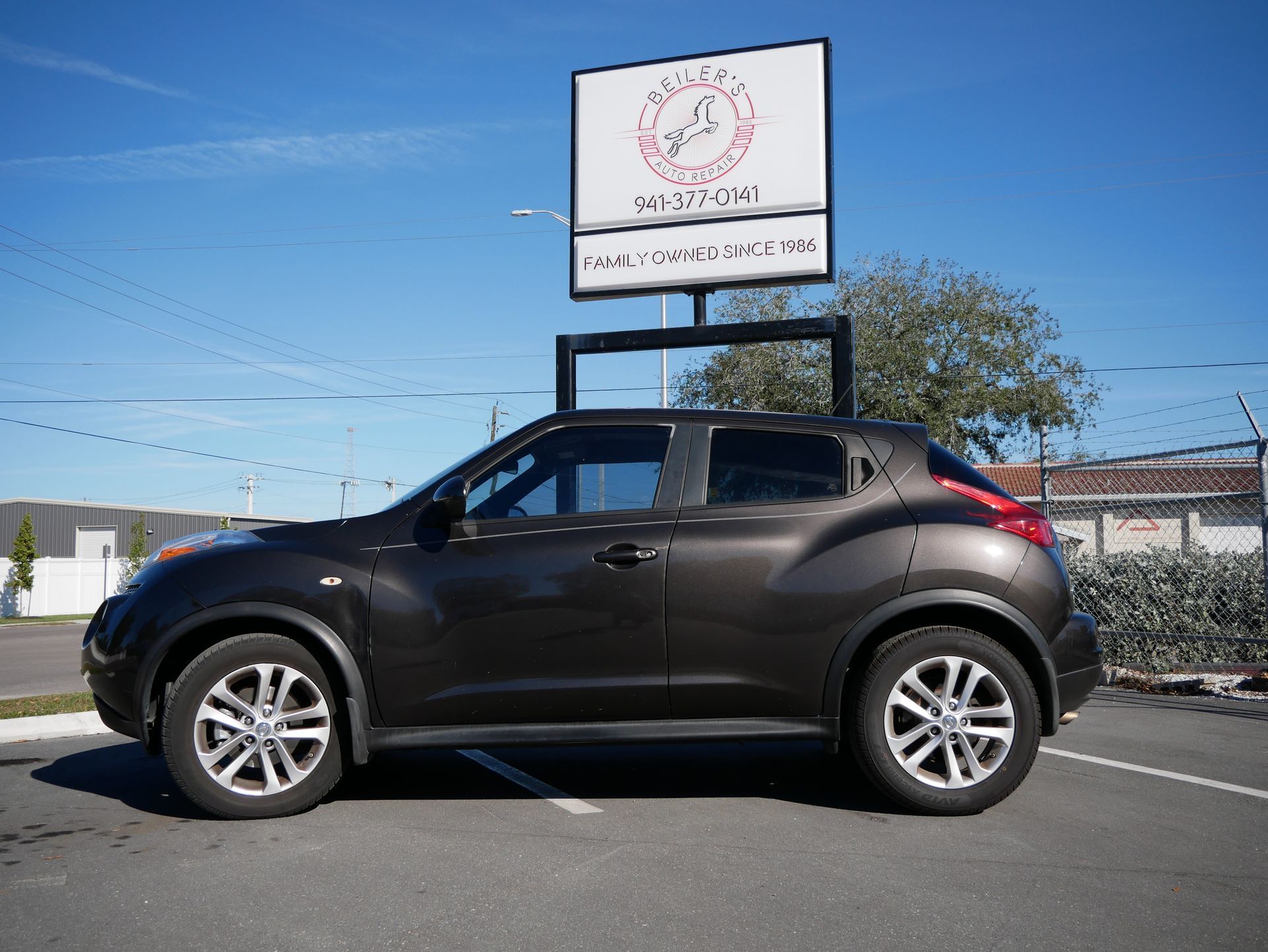 Dark gray Nissan Juke parked outside a business with a sign on a sunny day.