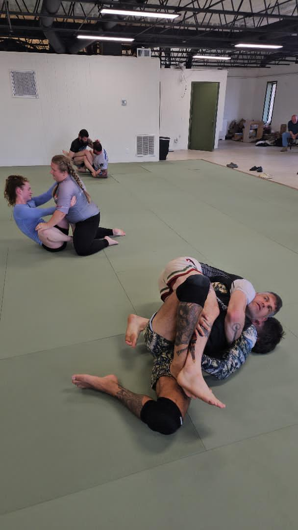Two people practicing grappling on mats, one in a gray hoodie pinning the other.