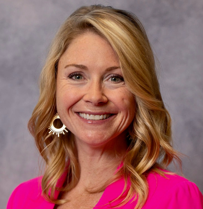 A woman wearing a pink jacket and earrings is smiling for the camera.