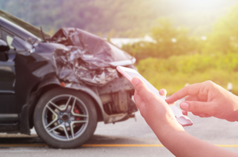 Picture of a black car has been in an accident. The front bumper is smashed in. You can see the hands of someone holding a phone indicating they are getting ready to call a Coquitlam Towing Company.