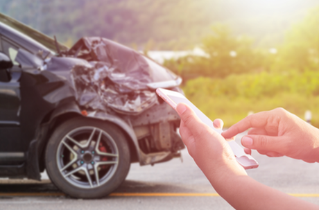 Picture of a black car has been in an accident. The front bumper is smashed in. You can see the hands of someone holding a phone indicating they are getting ready to call a Coquitlam Towing Company.