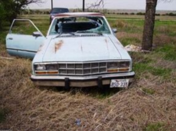Picture of a old scrap blue junk car. The car is sitting in a yard of grass.