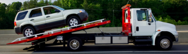 Picture of a white flatbed tow truck tilted. The tow truck is in the middle of towing on a silver vehicle.