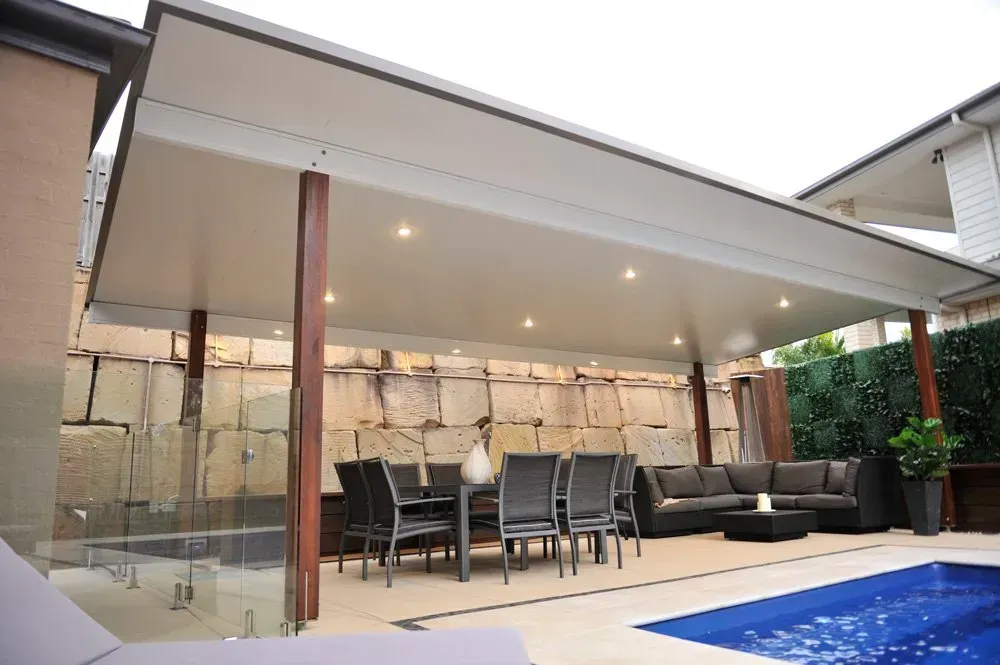 Patio with dining set, couches, and pool under a white roof with spotlights, supported by wooden posts.