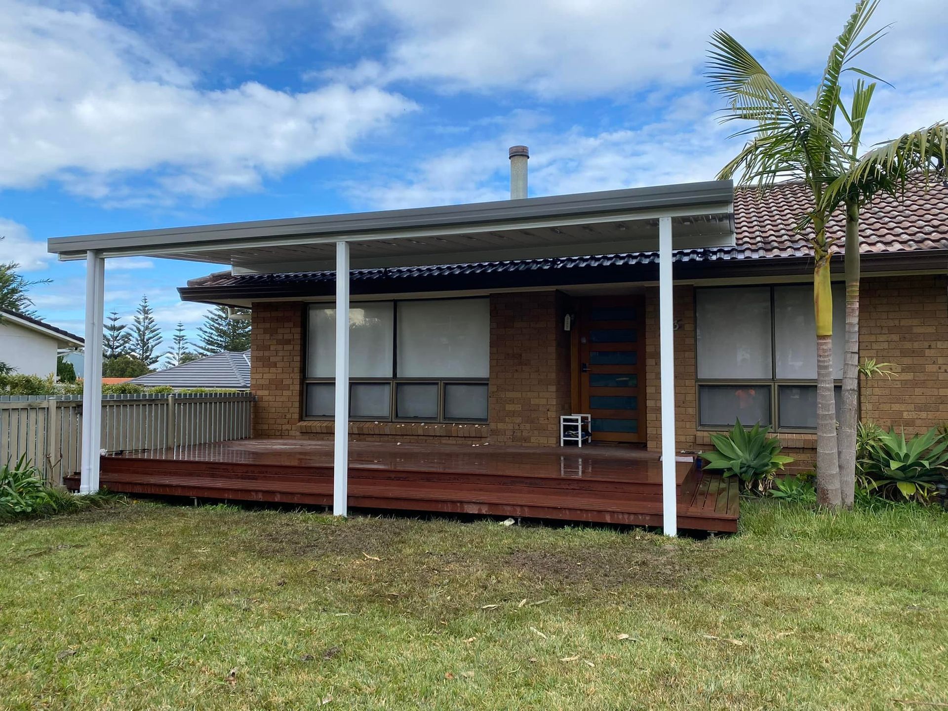 A brick house with a deck and awning, white pillars, a wooden deck, and grass in front.