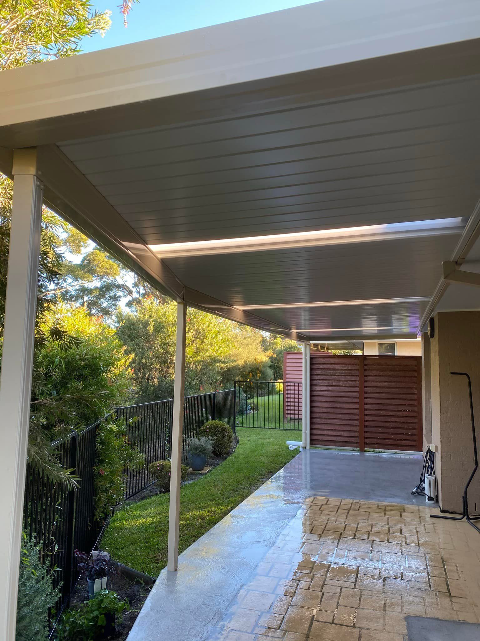 Covered outdoor patio with a view of a green yard, wooden fence, and paved area.