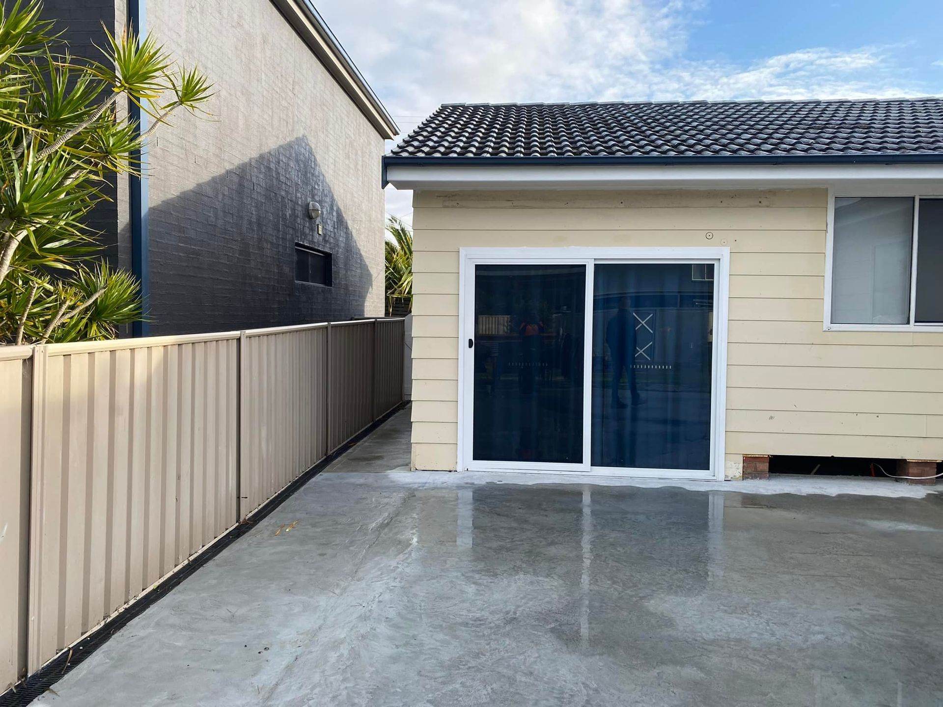 Beige building with sliding glass doors and a beige fence on a concrete surface under a cloudy sky.