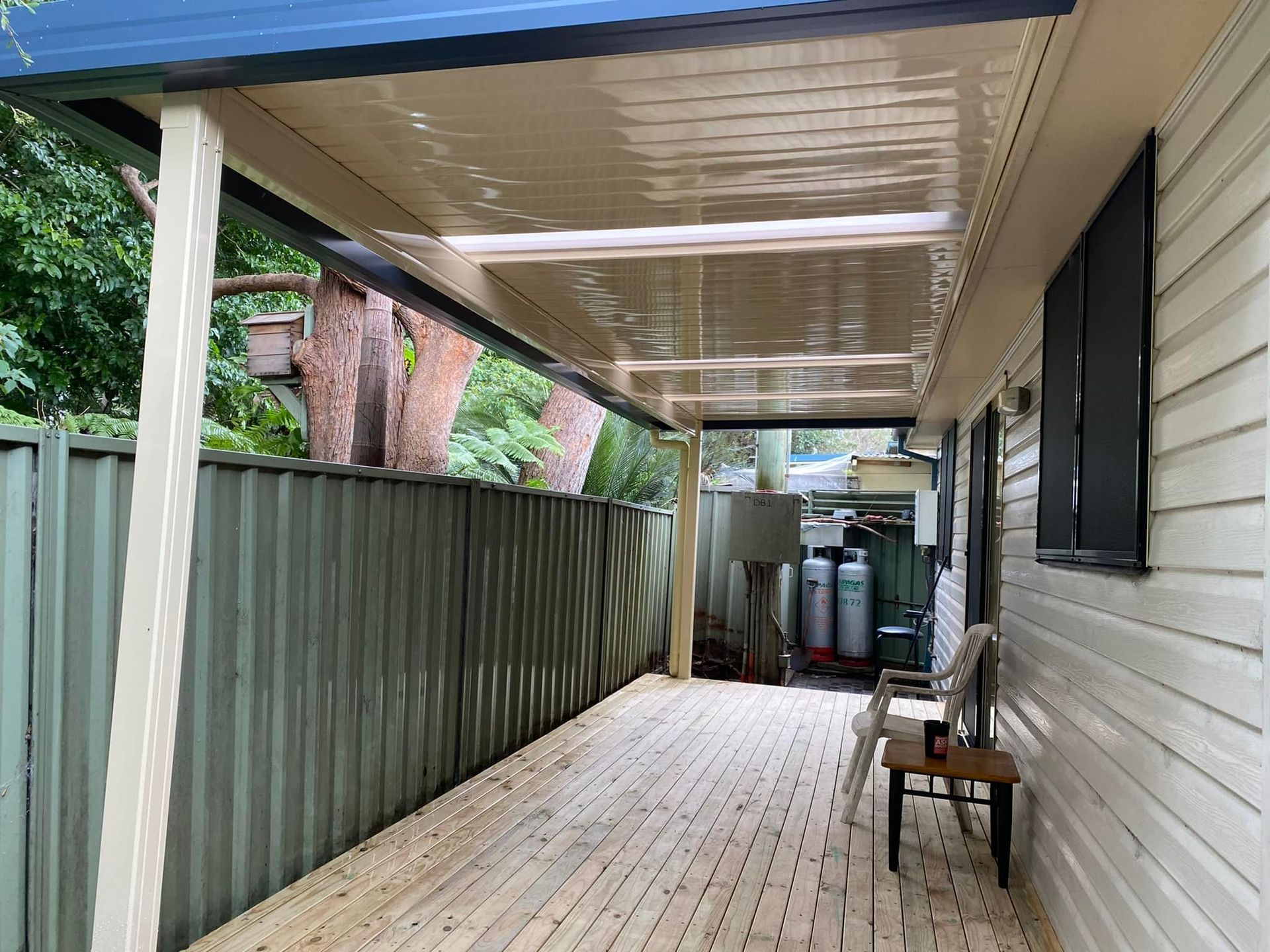 Covered outdoor deck with wooden floor, corrugated fence, and house wall.