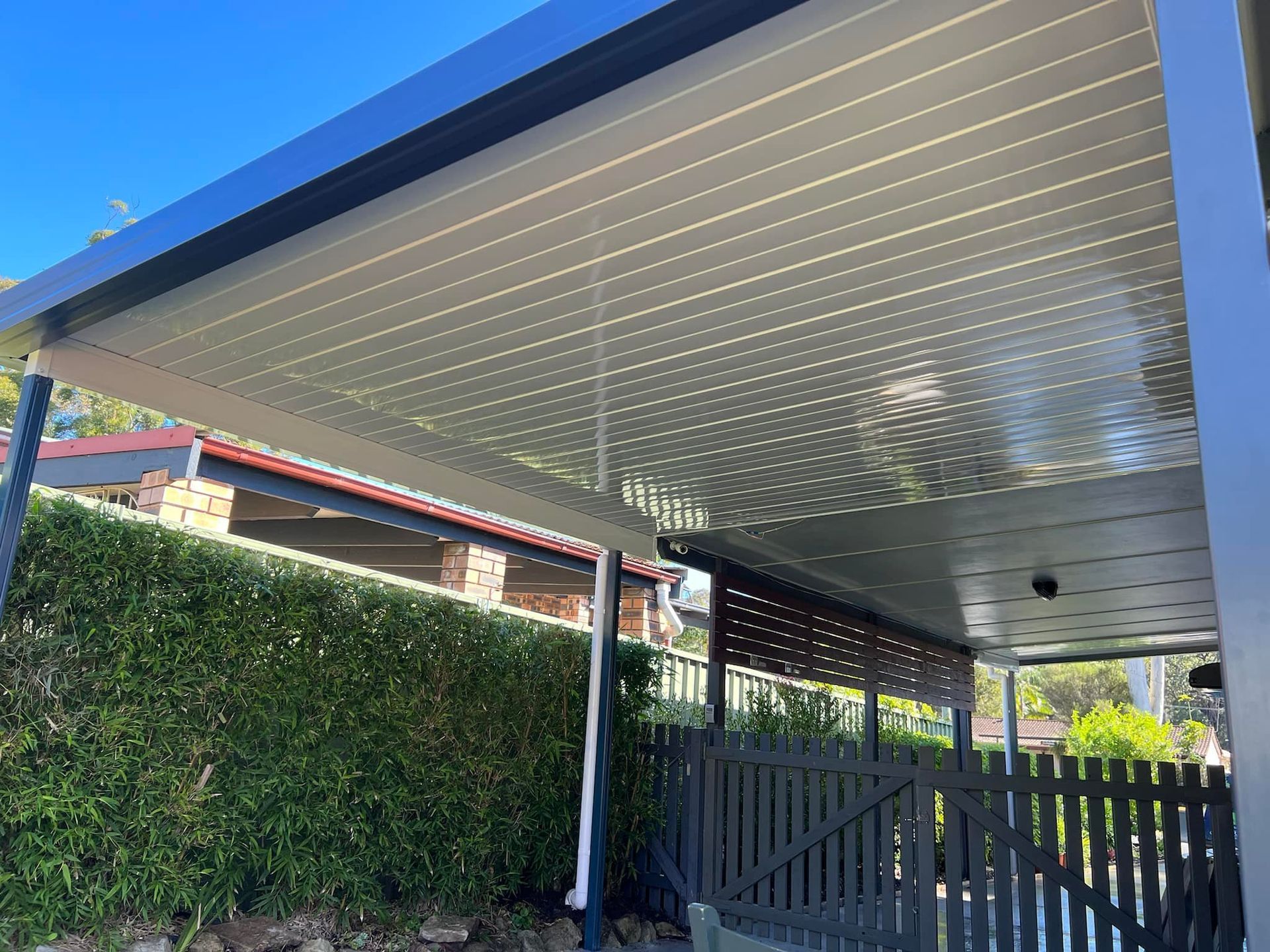 Patio roof with gray corrugated panels, dark gray frame, overlooking a garden and gate.