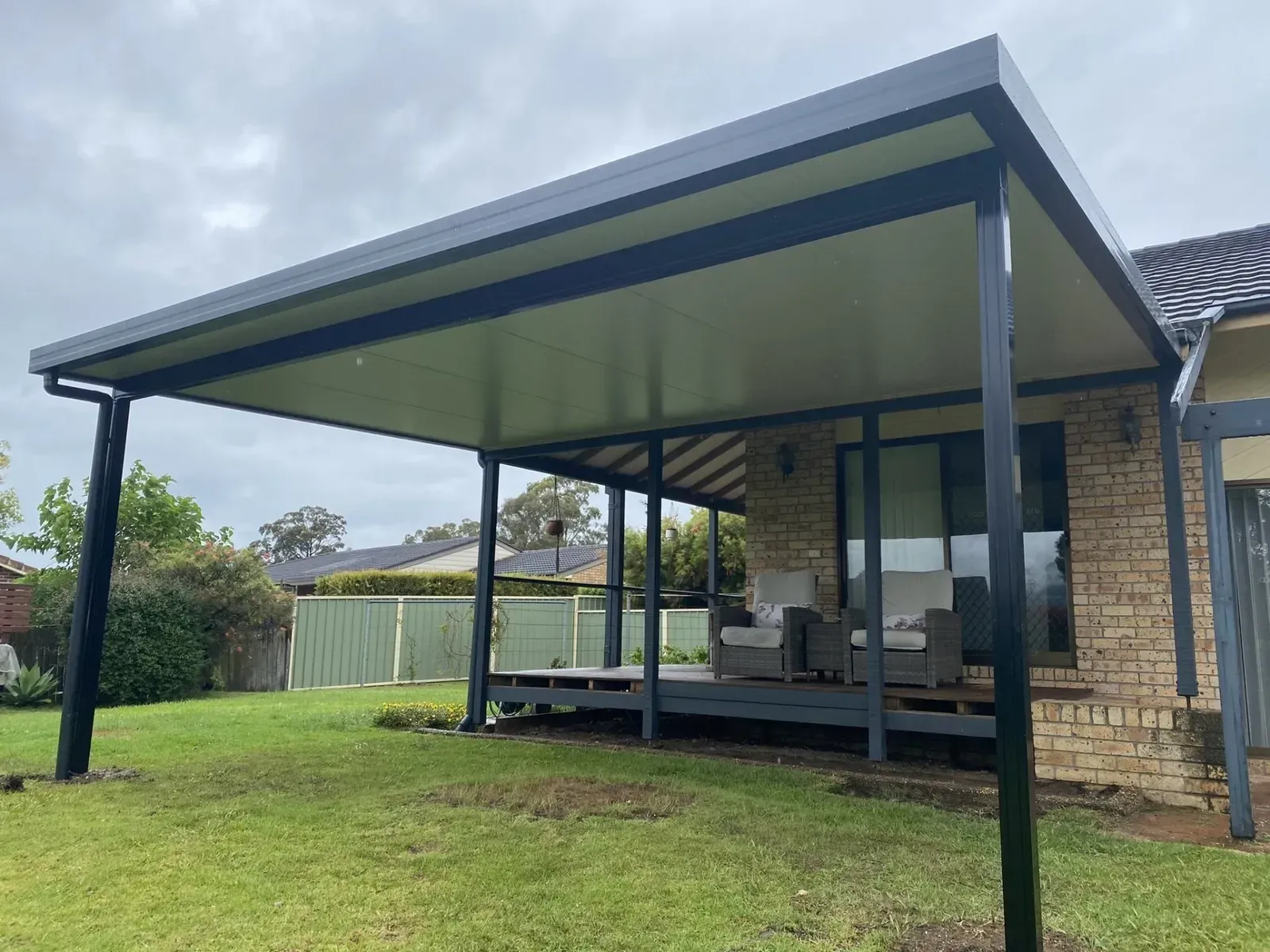 Patio cover attached to a brick house, supported by black posts, over a wooden deck with outdoor furniture.