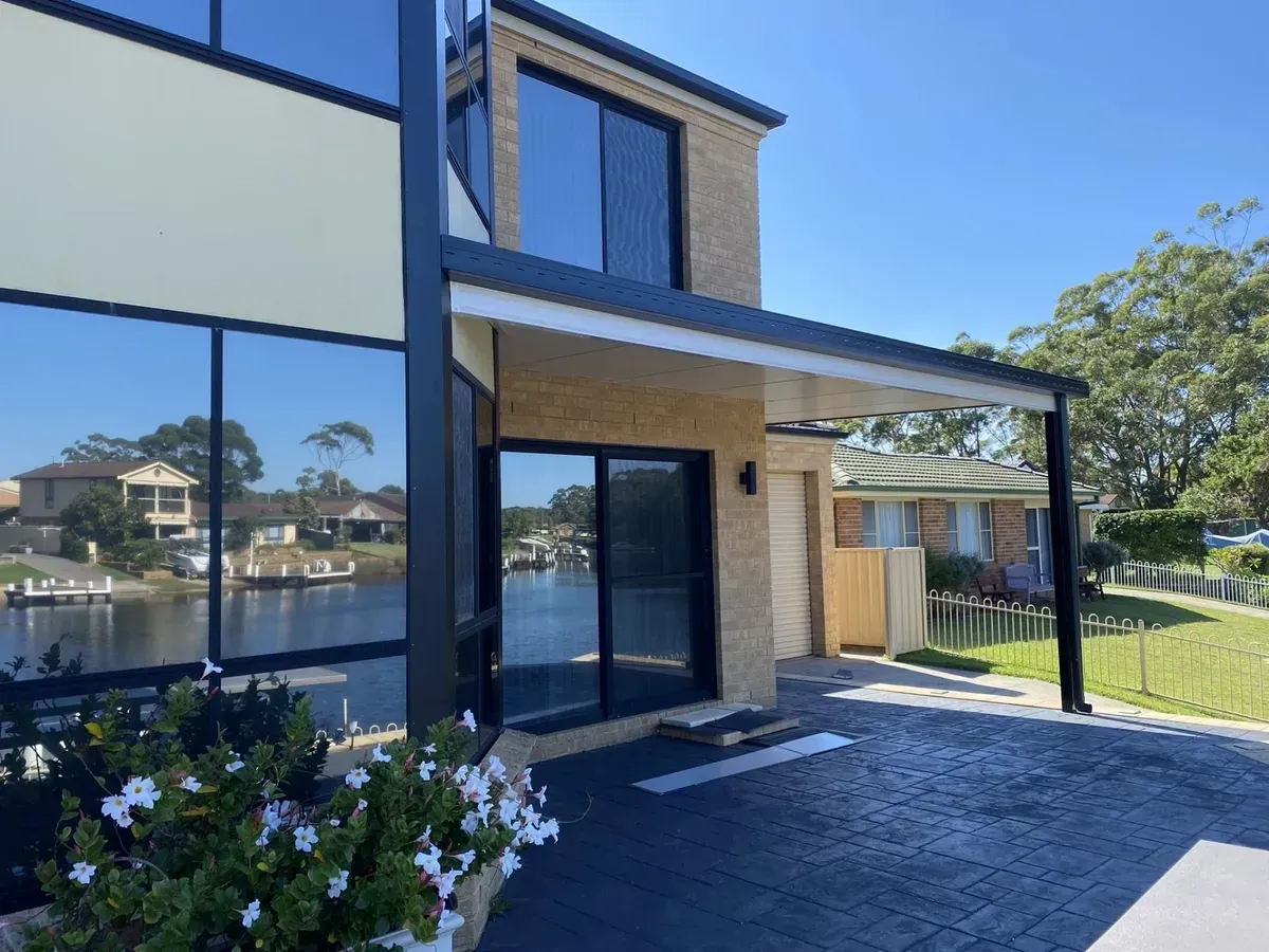 Modern two-story brick house with dark-tinted windows, awning, and water view on a sunny day.