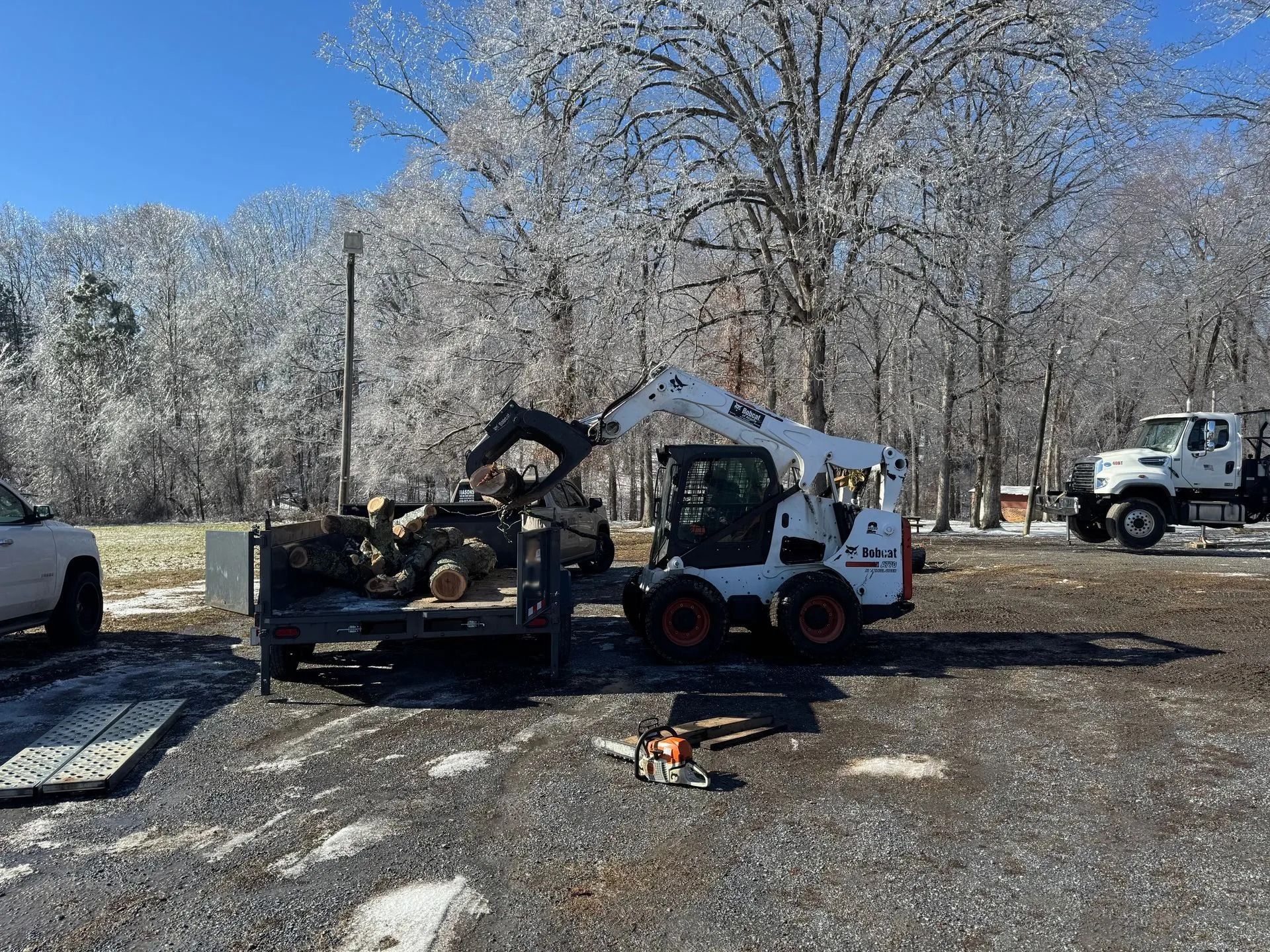 Bobcat loader with grapple clearing debris in a snowy yard, with a dump truck and work truck nearby.
