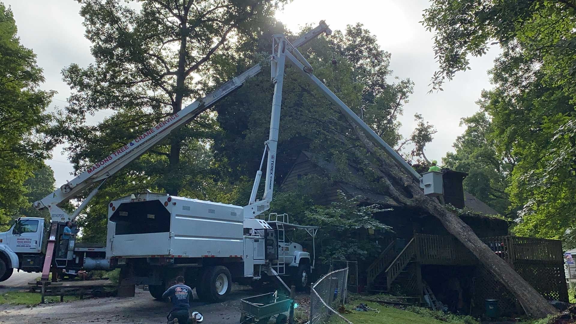 Two utility trucks support a collapsed A-frame roof amid trees after storm damage.