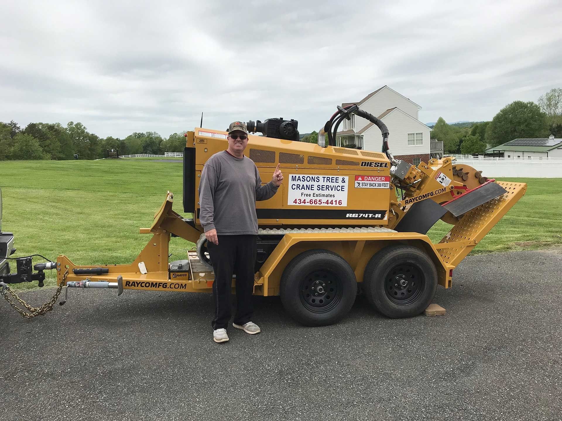 Man standing beside yellow wood chipper trailer on a gravel lot, with grassy field and cloudy sky behind.