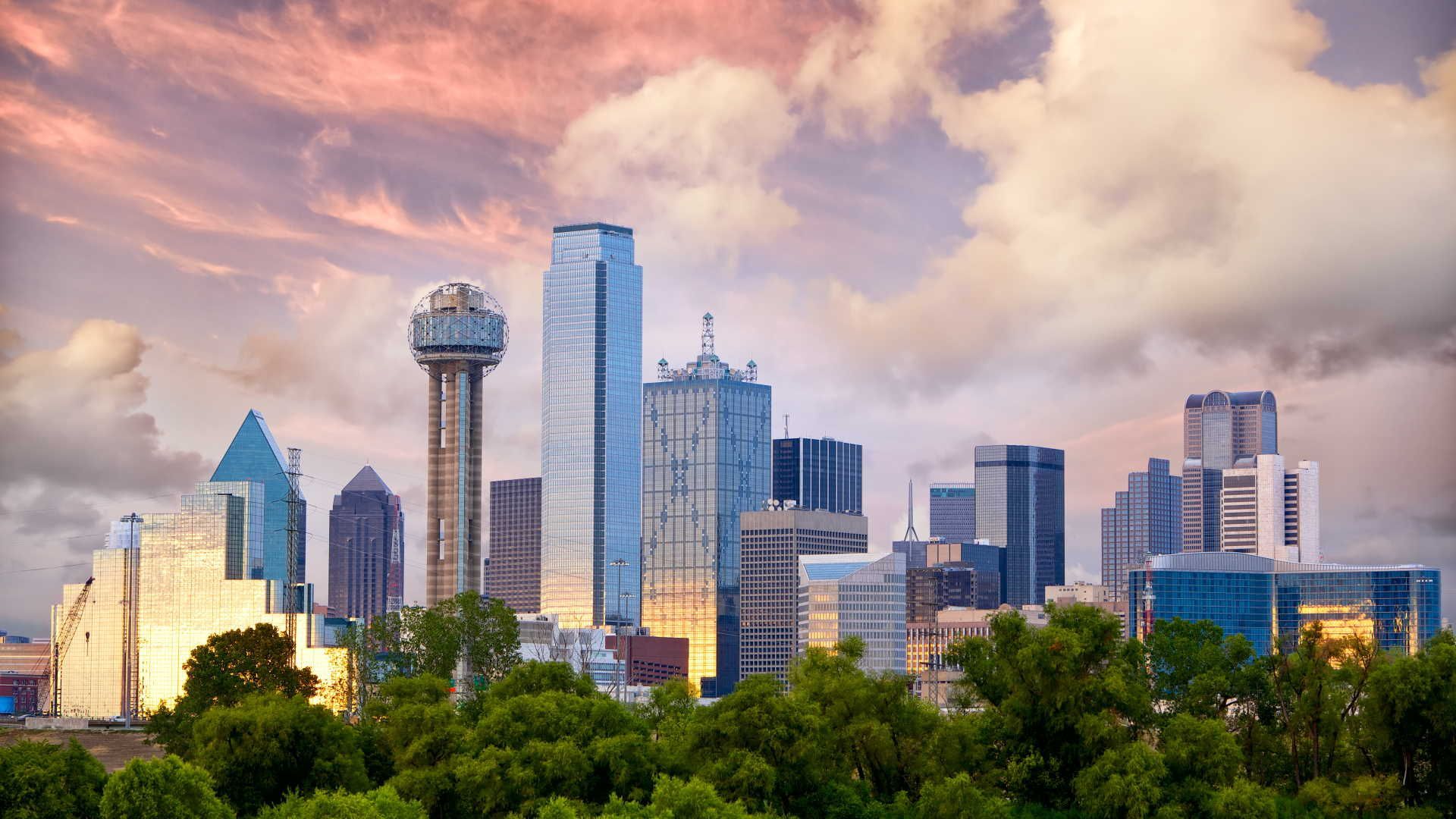 A city skyline with trees in the foreground and a cloudy sky in the background.