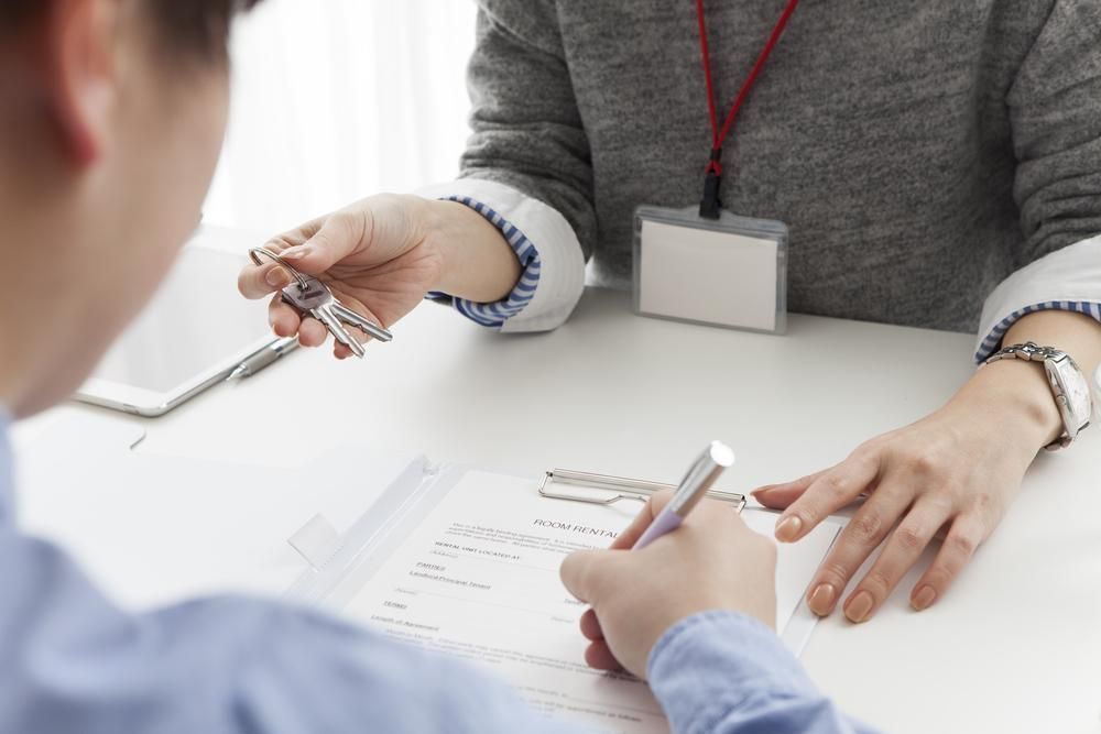 A Person Signing Paperwork at a Desk While Another Person Holds Keys, Likely a Real Estate Transaction 