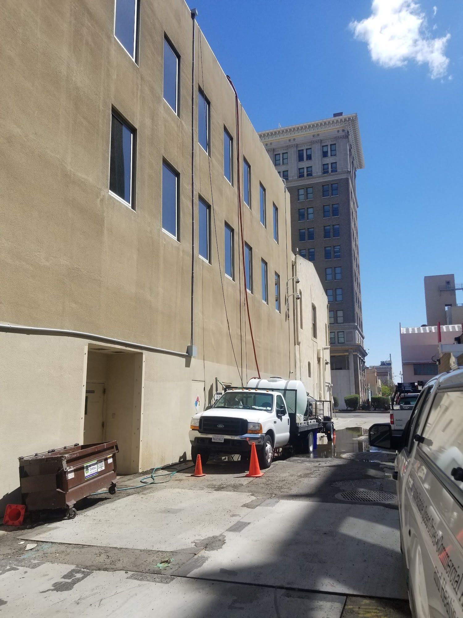Narrow city alley beside beige building with parked white van, cones, trash bin, and tall building in distance