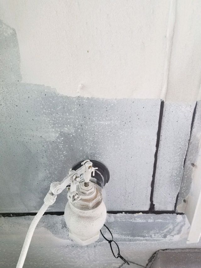 White wall with a small wall-mounted device, hose attached, and a black cord on the floor.