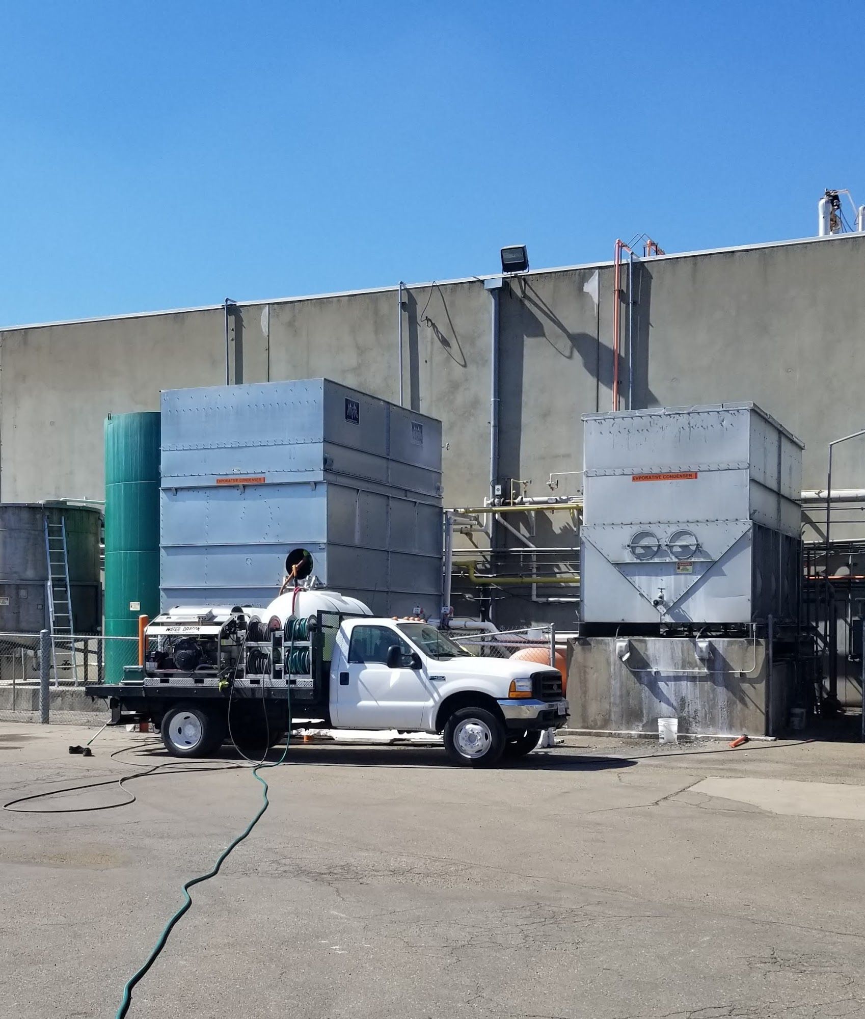 White pickup truck with water tank beside large concrete industrial tanks under a blue sky