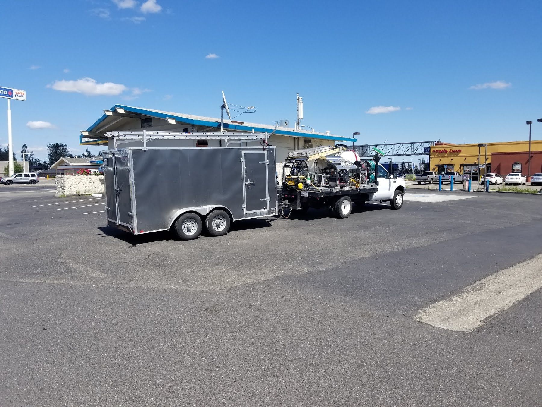 Gray trailer parked in an open lot beside a white truck under a blue sky