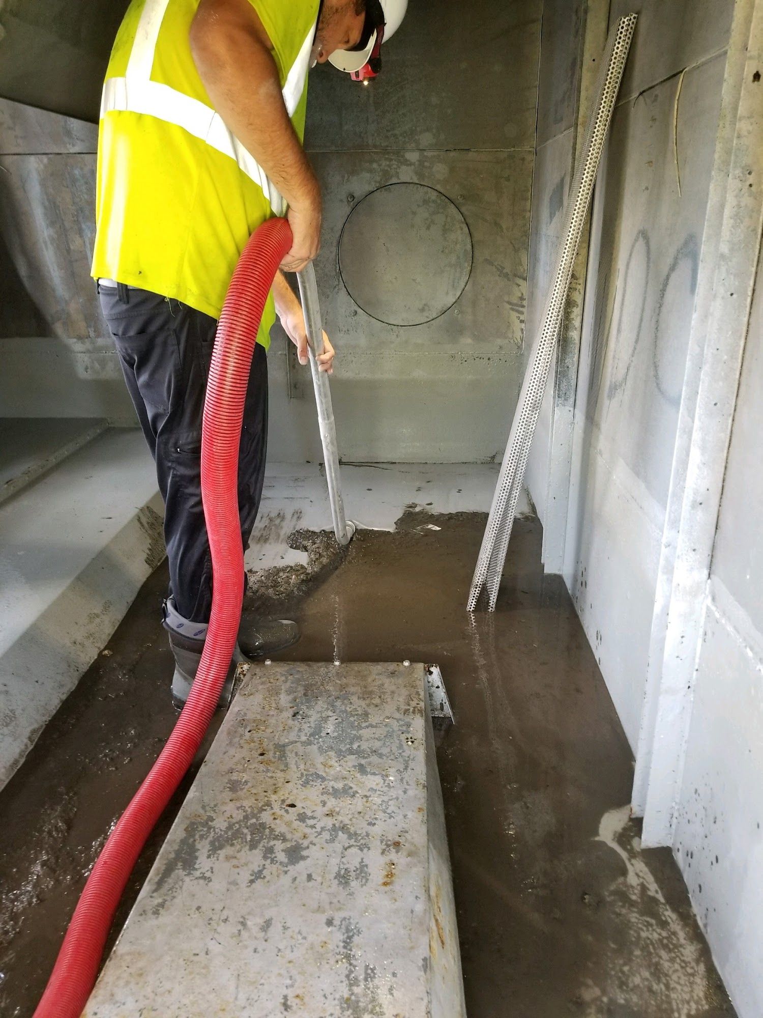 Worker in a yellow vest spraying water inside a concrete tank or utility chamber.