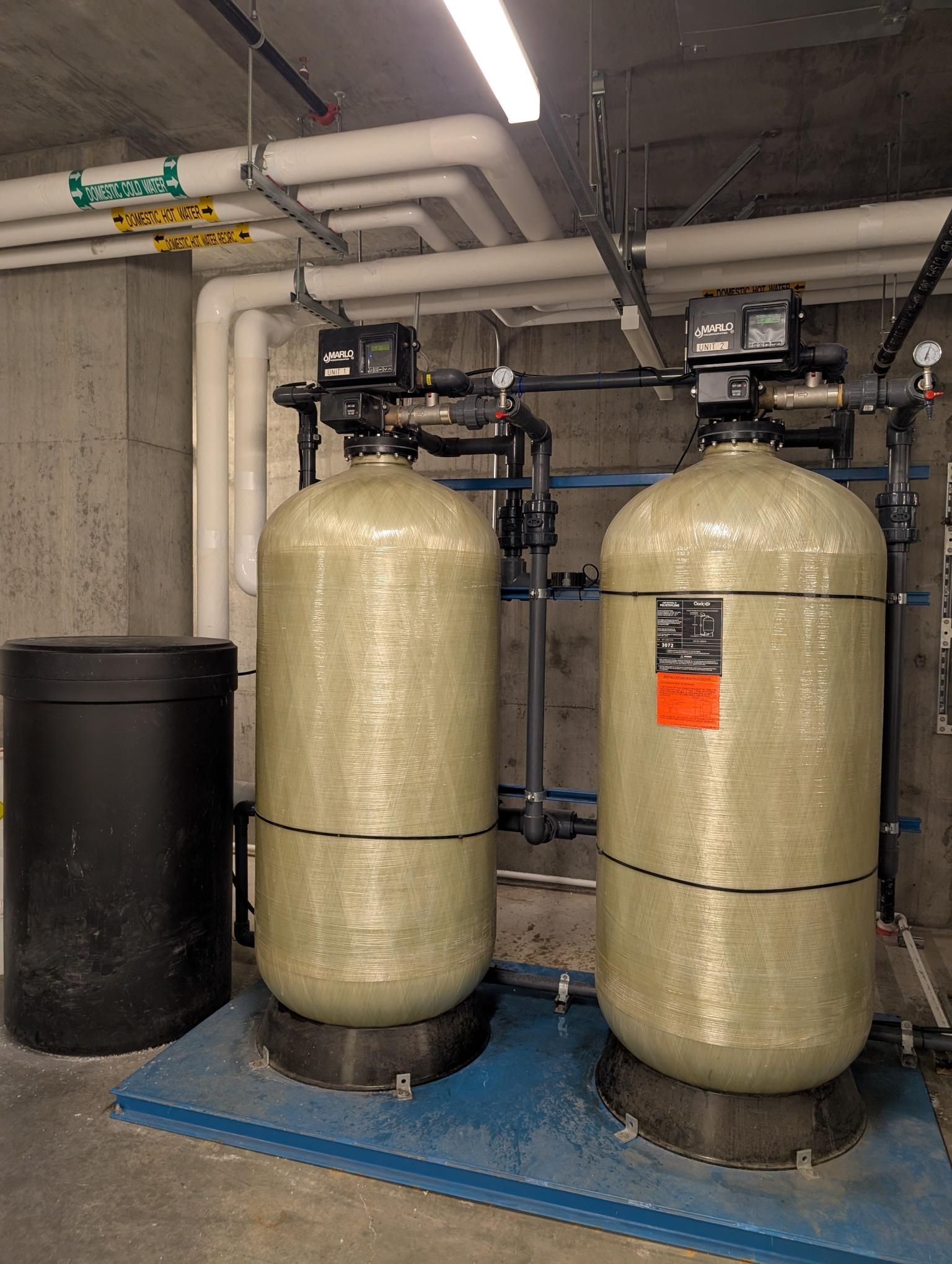 Two beige water softener tanks and a black tank in a basement utility room.