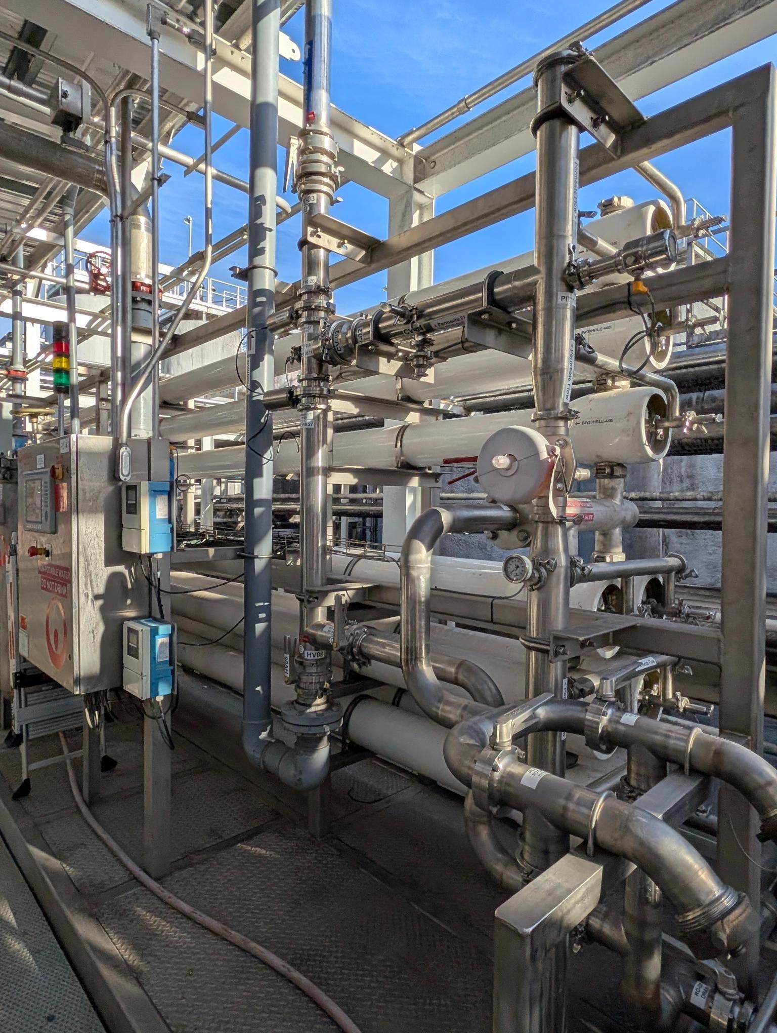 Industrial piping and metal machinery in a factory, with silver pipes, valves, and platforms under bright daylight