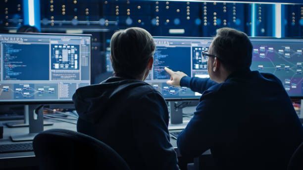 Two men are looking at computer monitors in a dark room.