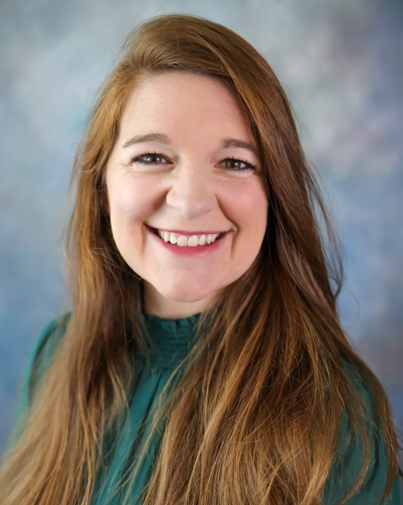 A woman with long brown hair is smiling and wearing a green shirt.