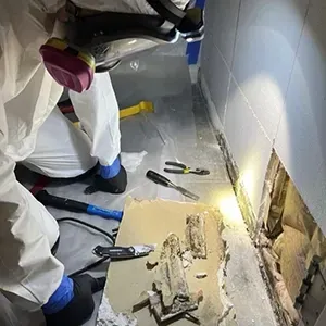 Workers in white protective suits inspect a damaged wall with debris on the floor.