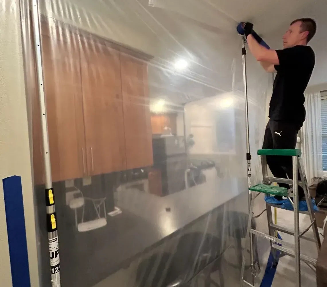 Worker on a ladder installing clear plastic sheeting over a large interior opening.