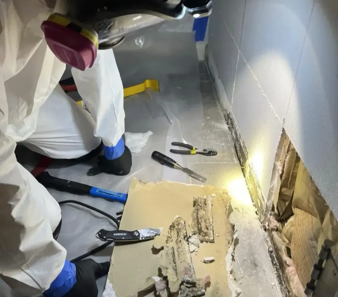 Workers in white protective suits inspect a damaged wall with debris on the floor.