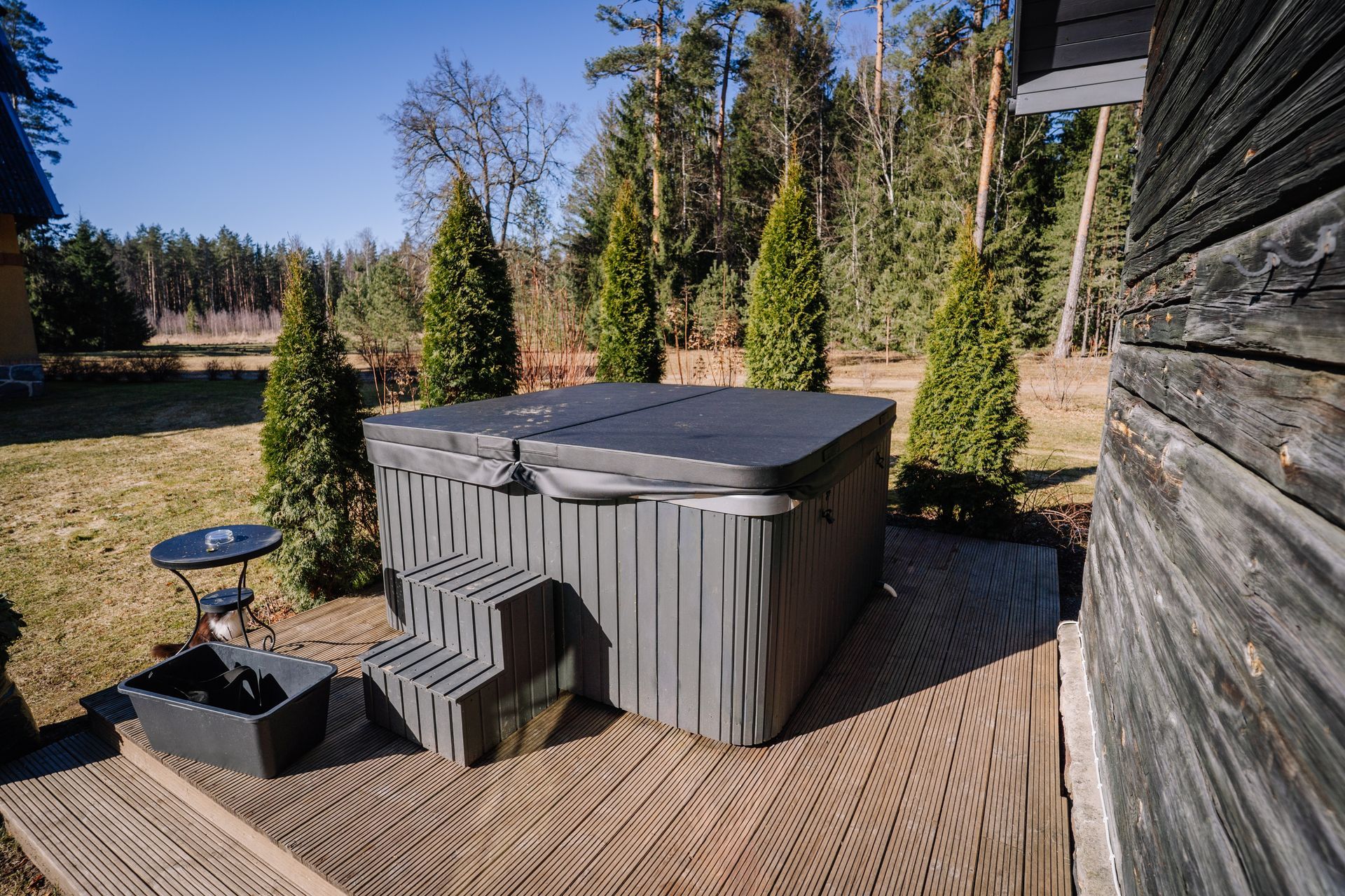 Backyard patio with a gray hot tub, small grill, and evergreen shrubs beside a wooden house