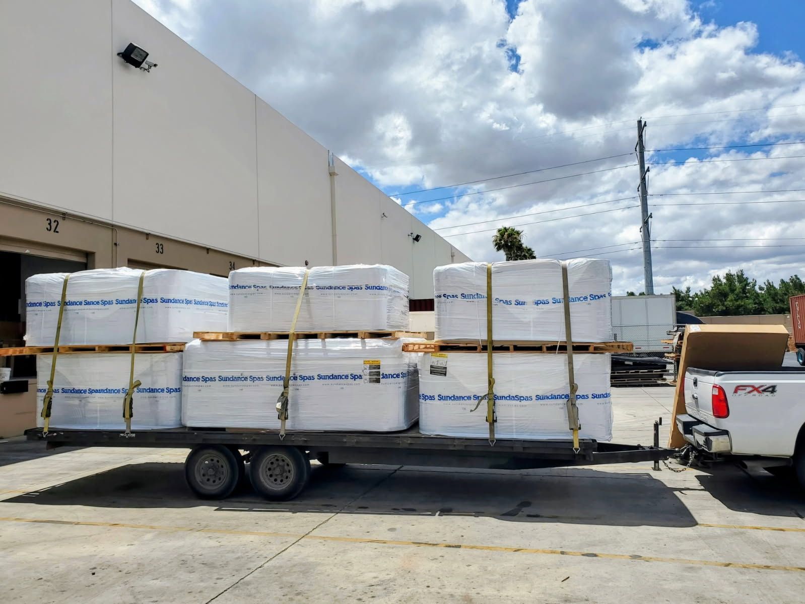 Flatbed trailer loaded with wrapped pallets beside a warehouse under a cloudy sky