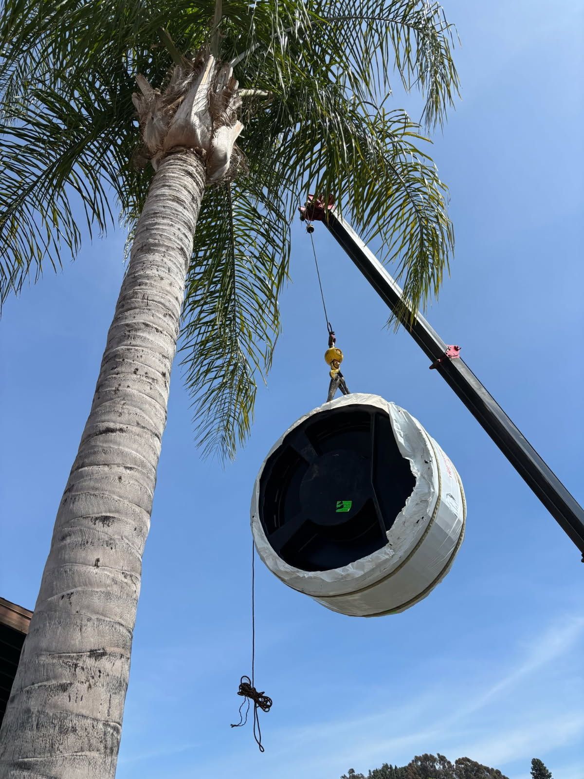 Crane hoists a large cylindrical duct section beside a palm tree under a blue sky.