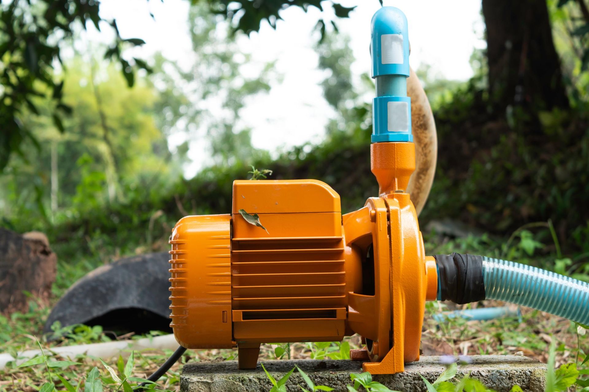 Orange water pump with blue pipe outdoors on grass, with a hose attached and trees in the background