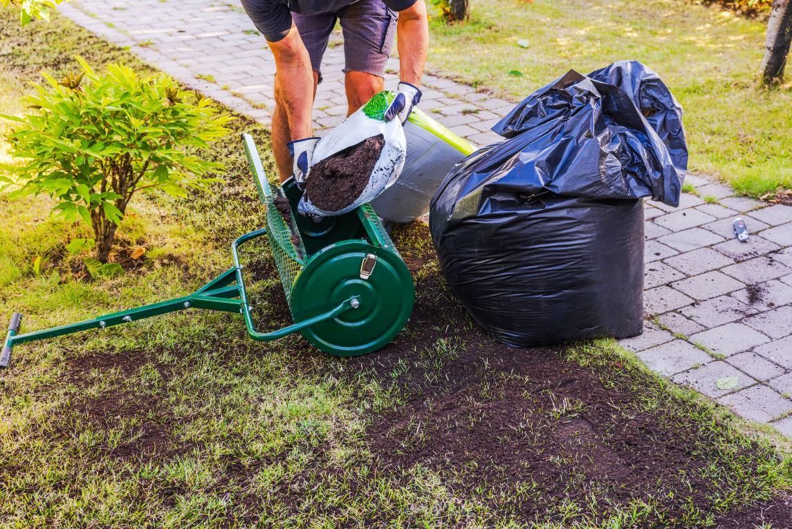 Person using a green lawn spreader beside a black trash bag on a grassy path