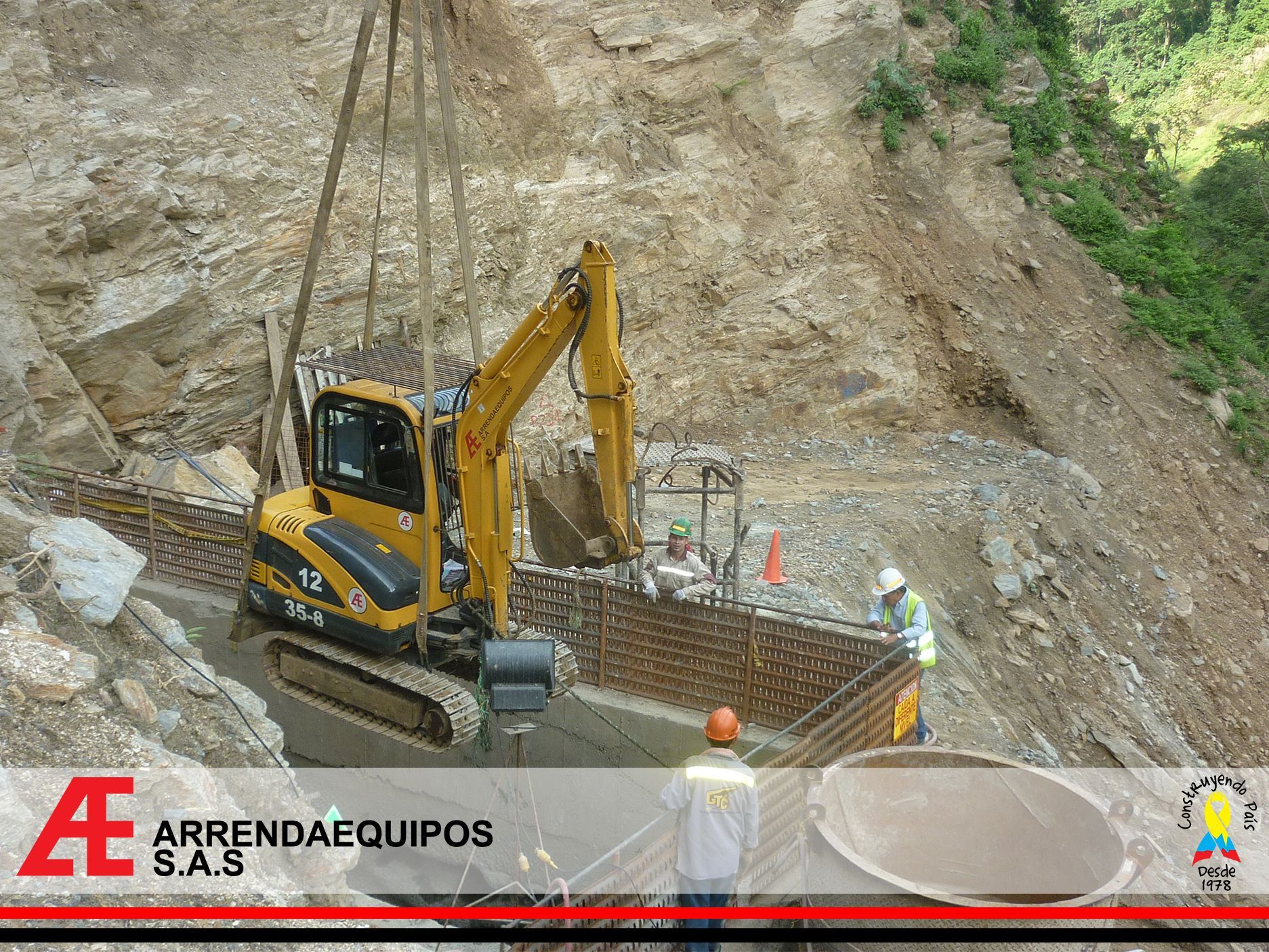 Excavadora amarilla y trabajadores construyendo un muro de contención a lo largo de una ladera rocosa.