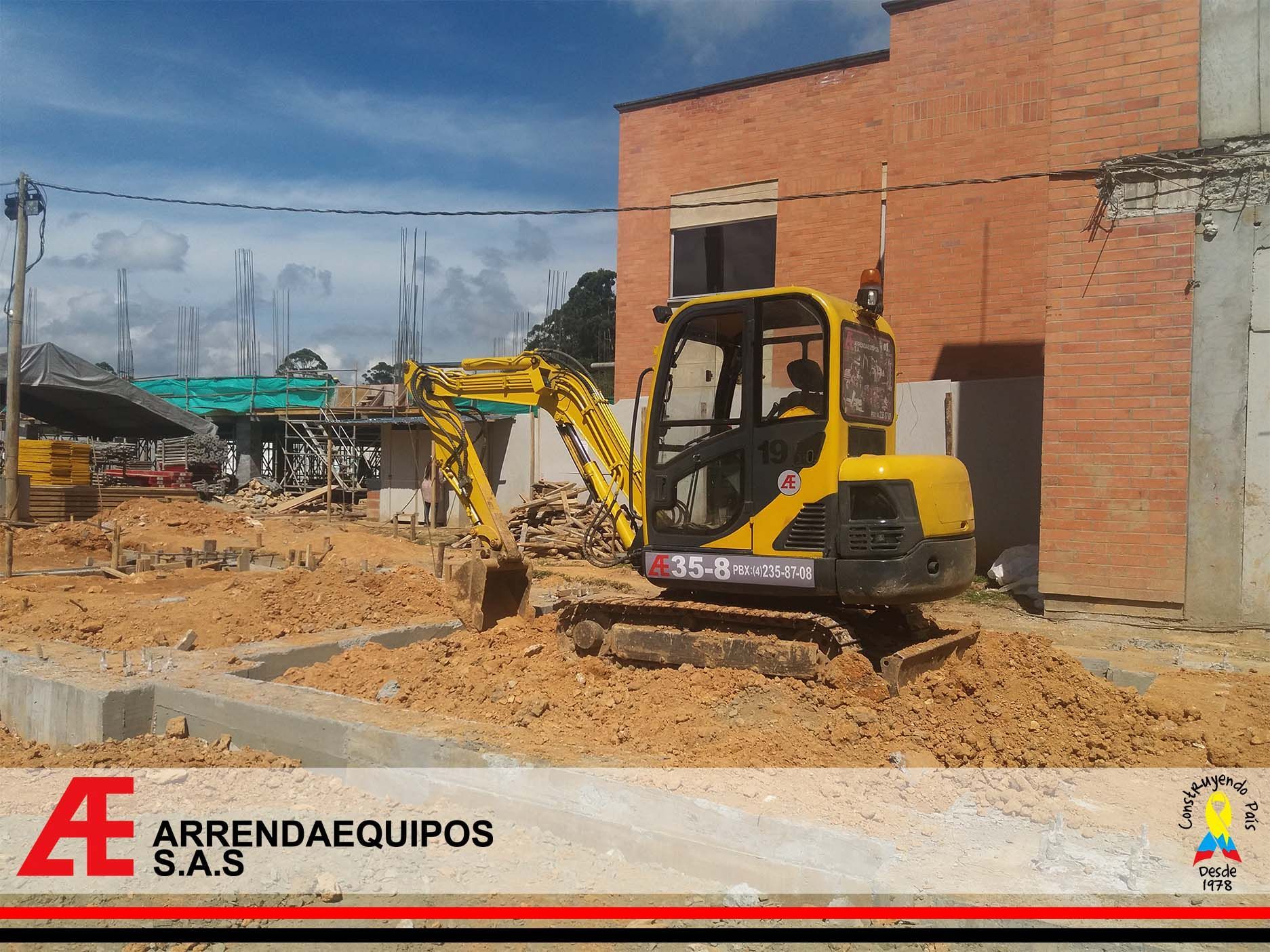 Excavadora amarilla en una obra, recogiendo tierra. Edificios y cielo azul al fondo.