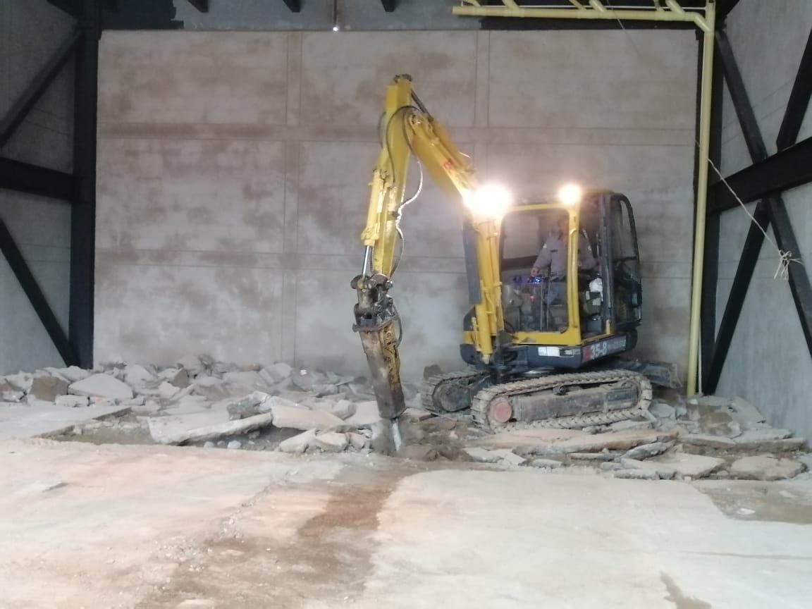 Una excavadora amarilla con un martillo neumático derriba el piso de concreto dentro de un edificio.