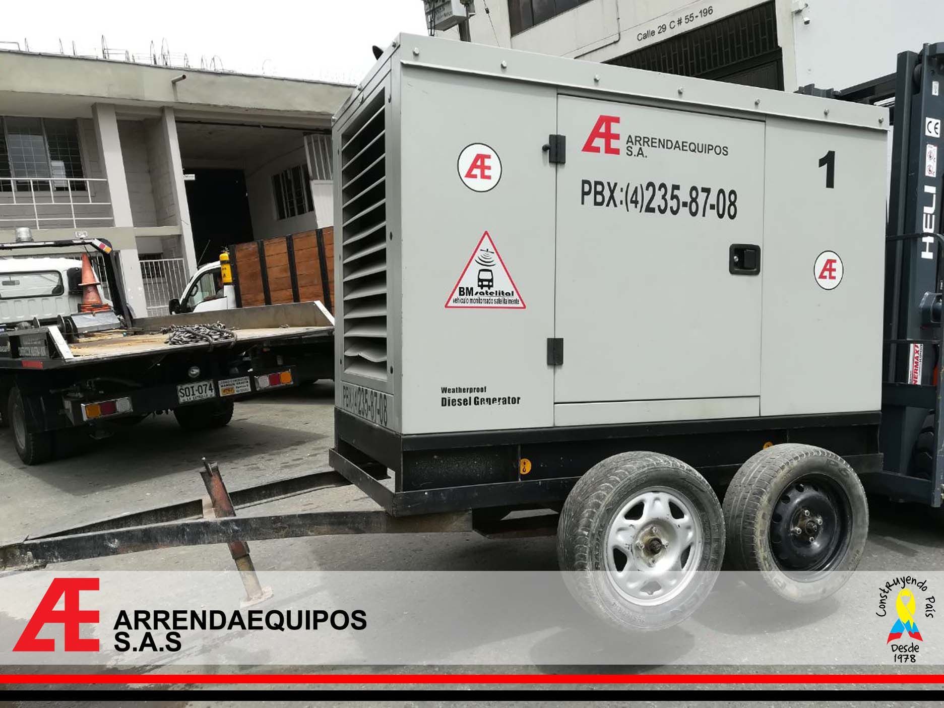 Generador sobre un remolque, gris, con marca, estacionado junto a un edificio. Se ven un camión y una carretilla elevadora.