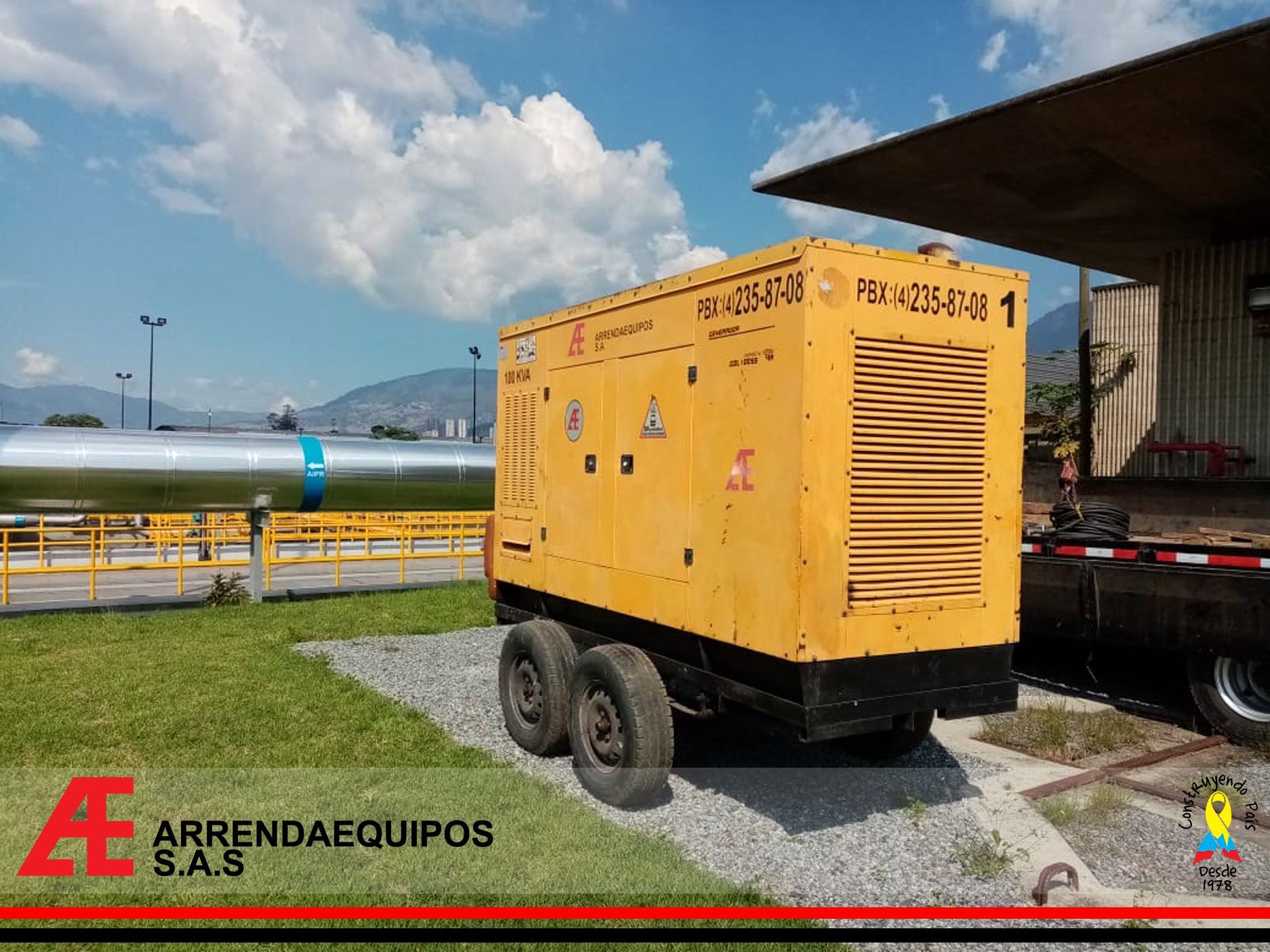 Generador amarillo sobre ruedas cerca de un edificio y una tubería en una zona cubierta de hierba, bajo un cielo nublado.