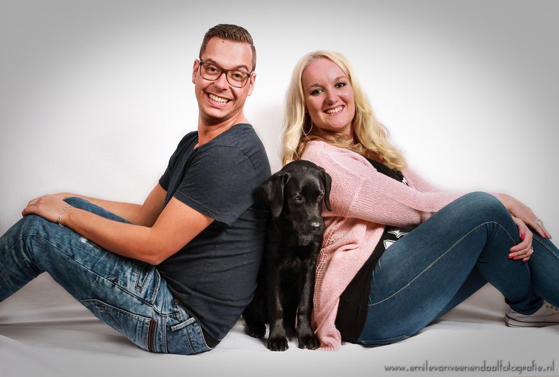 Twee mensen zitten rug aan rug met een zwarte hond tussen hen in, tegen een studioachtergrond.