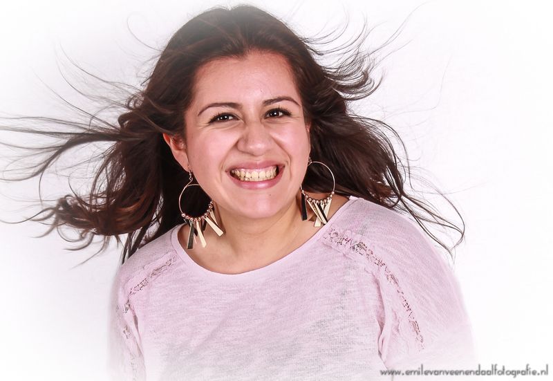 Lachende vrouw met wapperend haar, gekleed in een lichtroze trui, tegen een witte achtergrond.
