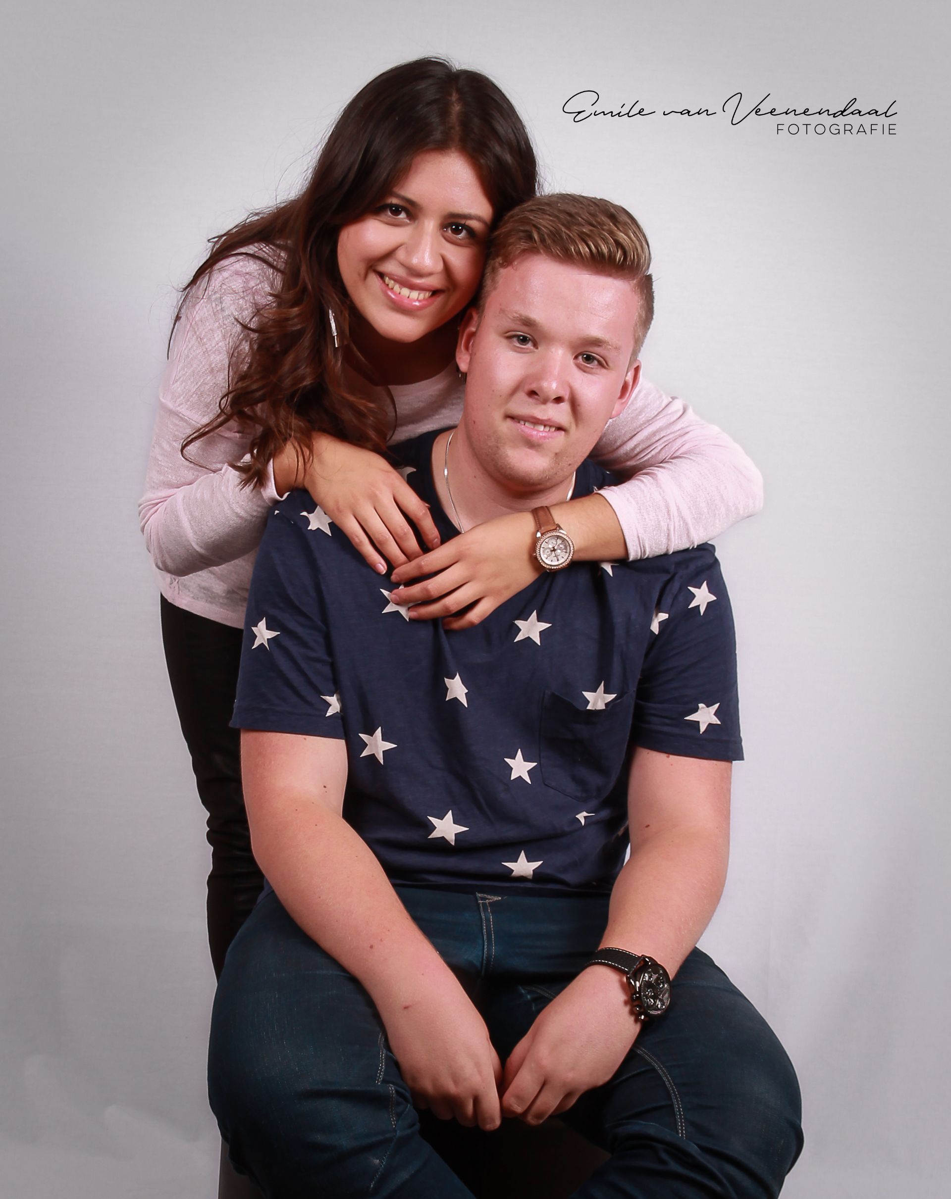Twee mensen poseren in een studio; de een staat achter de ander met de armen om hem heen, beiden glimlachend.